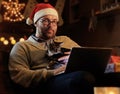 A man in Santa`s New Year hat holds a cat and working with laptop. Royalty Free Stock Photo