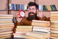 Man, scientist peeking out of piles of books with alarm clock. Teacher with beard studying in library. Time flow concept