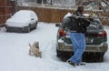 Man scraping ice from car with two Westie dogs looking on in heavy snow