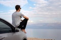 Man seated on the engine hood of a rented car on a road trip in israel