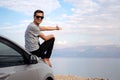 Man seated on the engine hood of a rented car on a road trip in israel