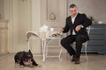 Man seatting at the table and looking at the watch. Near leas a dog pit bull or black Staffordshire Terrier in vintage interior