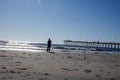 A man seen standing at the ocean water edge holding his fishing rod on an empty beach with a long pier in the background Royalty Free Stock Photo