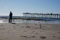 A man seen standing at the ocean water edge holding his fishing rod on an empty beach with a long pier in the background Royalty Free Stock Photo