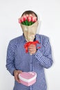 A man in a shirt holds tulips flowers in front of his face and holds out a heart-shaped box on a white background. Royalty Free Stock Photo