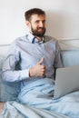 A man in a shirt sits on a bed and works on a laptop