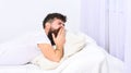 Man in shirt yawning while laying on bed, white wall and curtain on background. Sleepyhead concept. Macho with beard and