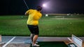 A man with a short pants holding a golf club playing at the golf driving range on a green carpet mat during the night