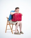 Man in shorts and a t-shirt with a red suitcase sits on a chair waiting, isolated on light background