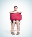 Man in shorts and a t-shirt with a red suitcase sits on a chair waiting, isolated on light background. Front view.