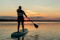 A man in shorts and a T-shirt on a SUP board with an oar against the backdrop of the sunset sky swim in the lake. Royalty Free Stock Photo