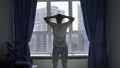 Man silhouette watching through window, stretching arms behind the head. Media. Man by the window with blue curtains.