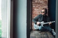 Man singer sitting on a window sill in a headphones with a guitar recording a track in a home studio Royalty Free Stock Photo