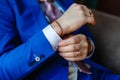 The man sits in an armchair and fixes the cuffs on a white shirt and a stylish blue jacket with expensive gold accessories, watche