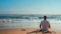 A man sits crosslegged on the sand facing towards the endless stretch of ocean back to the camera. closed eyes and