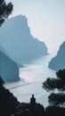 Man sits on a rocky outcrop overlooking a calm sea inlet, flanked by towering cliffs Royalty Free Stock Photo