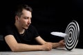 Man sits at table holding dart board with quill pen stuck in the center of the target on black background Royalty Free Stock Photo