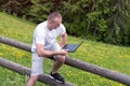 Man sits on a wooden fence and works with a laptop near the field and coniferous forest Royalty Free Stock Photo