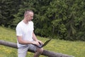 Man sits on a wooden fence and works with a laptop near the field and coniferous forest Royalty Free Stock Photo