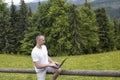 Man sits on a wooden fence and works with a laptop near the field and coniferous forest Royalty Free Stock Photo