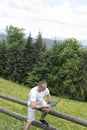 Man sits on a wooden fence and works with a laptop near the field and coniferous forest. Vertical frame Royalty Free Stock Photo