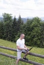 Man sits on a wooden fence and works with a laptop near the field and coniferous forest. Vertical frame Royalty Free Stock Photo