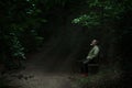 Man sitting on a bench in light spot on forest path, surrounded with darkness, looking up full of hope Royalty Free Stock Photo