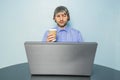 Man sitting by a computer holding cup with coffee. Model with grey hair and beard in his 40s wearing blue shirt. Working in an