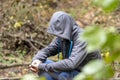 A man is sitting in a jacket with a hood hiding his face in the forest under a tree. Perhaps he is a lost tourist, a fugitive, an