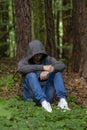 A man is sitting in a jacket with a hood hiding his face in the forest under a tree. Perhaps he is a lost tourist, a fugitive, an