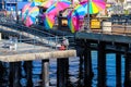 A man sitting on the long brown wooden pier fishing surrounded by colorful umbrellas with people walking along the pier