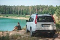 Man sitting near white suv car at the edge looking at lake with blue water