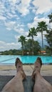 Man sitting relax in beautiful pool side showing only feet swiming pool palm tree blue cloudy skies and mountain as background Royalty Free Stock Photo