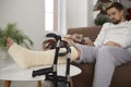 Man sitting on the sofa with his broken leg resting on a foot stool, and crutches leaning on it Royalty Free Stock Photo
