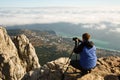 Man sitting with a tripod and photo camera on a high mountain peak above clouds, city and sea. Pro photographer Royalty Free Stock Photo