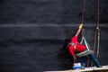 Man sitting on a wooden suspended scaffold and painting a wall in the color black