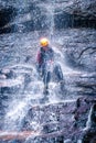 Man slips and gets wet abseiling down slippery rock face under a cascading waterfall hanging from a rope in wetsuit, helmet and