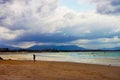 Man slumping - tired or sad - walking on beach coming out of surf at golden hour with beach full of people and mountians and storm