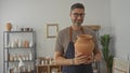 Man smiling and holding a terracotta jar with both hands in studio surrounded by pottery shelves and tools; calm contentment Royalty Free Stock Photo