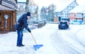 Man with snow shovel cleans sidewalks in winter during snowfall. Winter time in Europe. Young man in warm winter clothes