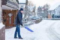 Man with snow shovel cleans sidewalks in winter during snowfall. Winter time in Europe. Young man in warm winter clothes