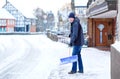 Man with snow shovel cleans sidewalks in winter during snowfall. Winter time in Europe. Young man in warm winter clothes