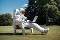 A man in a space suit sits on a lawn chair and works on a laptop