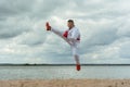 A man in a sports kimono with a red belt fulfills a karate jump kick. Training takes place on the beach.