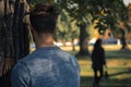 Man stalking woman while hiding behind tree in public park with blurry woman in background Royalty Free Stock Photo