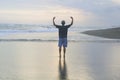 Man standing alone from behind on the beach, with arm raised against the sea at sunset.