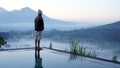 Man standing alone from behind on the edge of the pool at sunrise with water reflection and blue mount Batur view background.