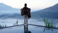 Man standing alone from behind on the edge of the pool at sunrise with water reflection and blue mount Batur view background.