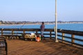 A man standing on the edge of a brown wooden pier leaning on the railing fishing with vast deep blue ocean water