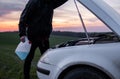 Man standing near car with open hood and holding a bottle with blue fluid on sunset. Car breakdown. Confused man stands